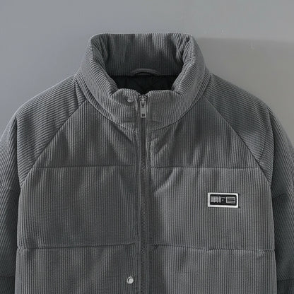 A gray ribbed corduroy puffer jacket with a stand-up collar, front zipper, and a small rectangular logo patch on the left chest, shown against a plain gray background.