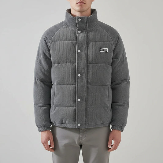A man wearing a gray quilted puffer jacket with a stand-up collar and snap-button front, paired with light gray trousers, against a plain light gray studio background.