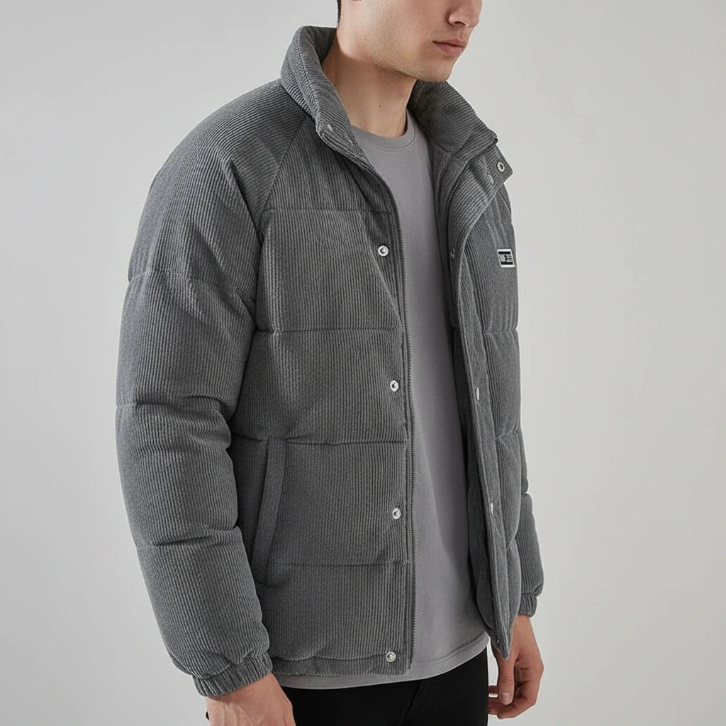 A man wearing a gray corduroy puffer jacket over a matching gray t-shirt, shown from the chest down against a light gray studio background.
